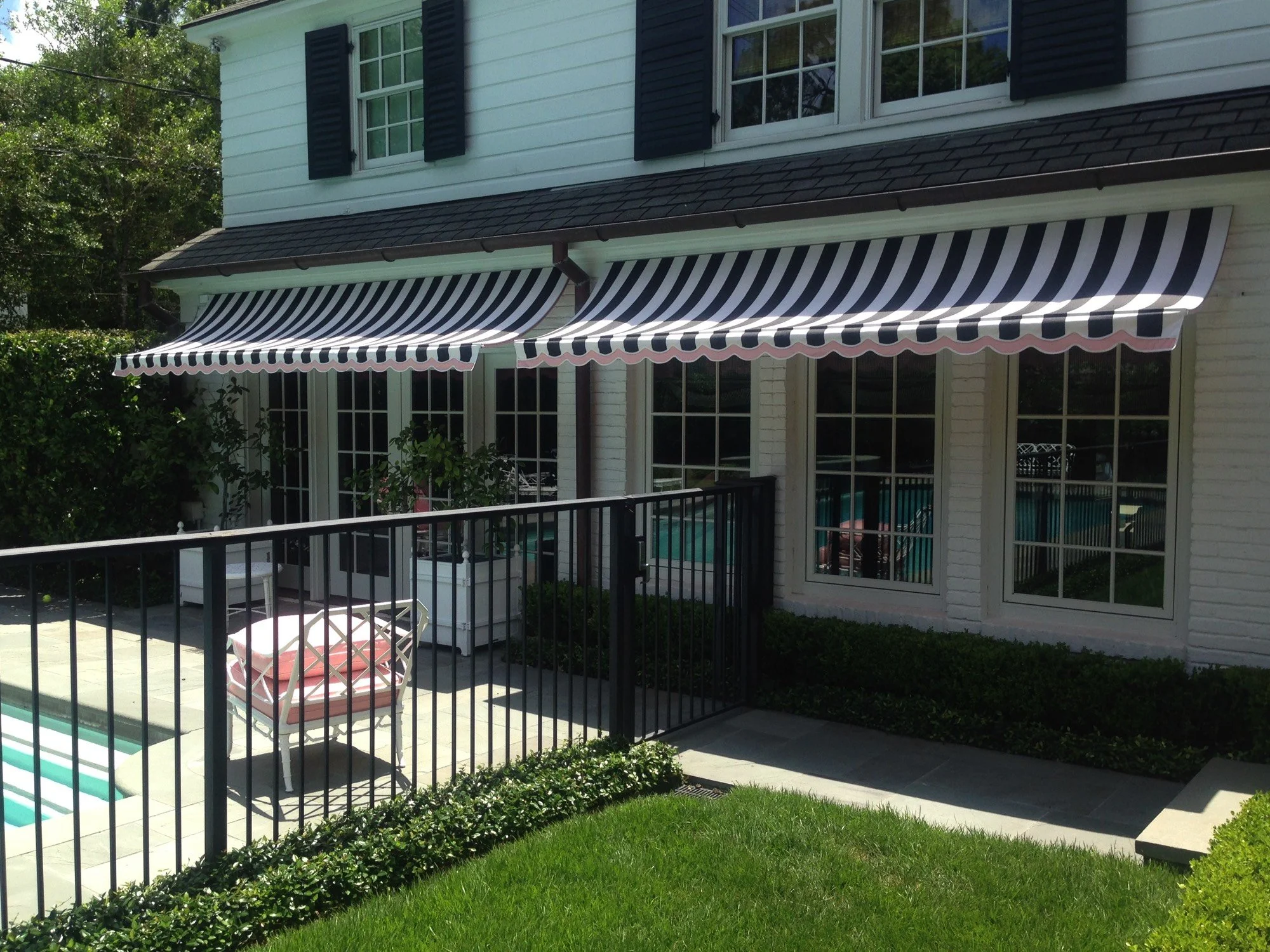 Backyard patio with black metal railing, white patio furniture with pink cushions, green grass, and striped black and white awnings over sliding glass doors attached to a white house with black shutters.