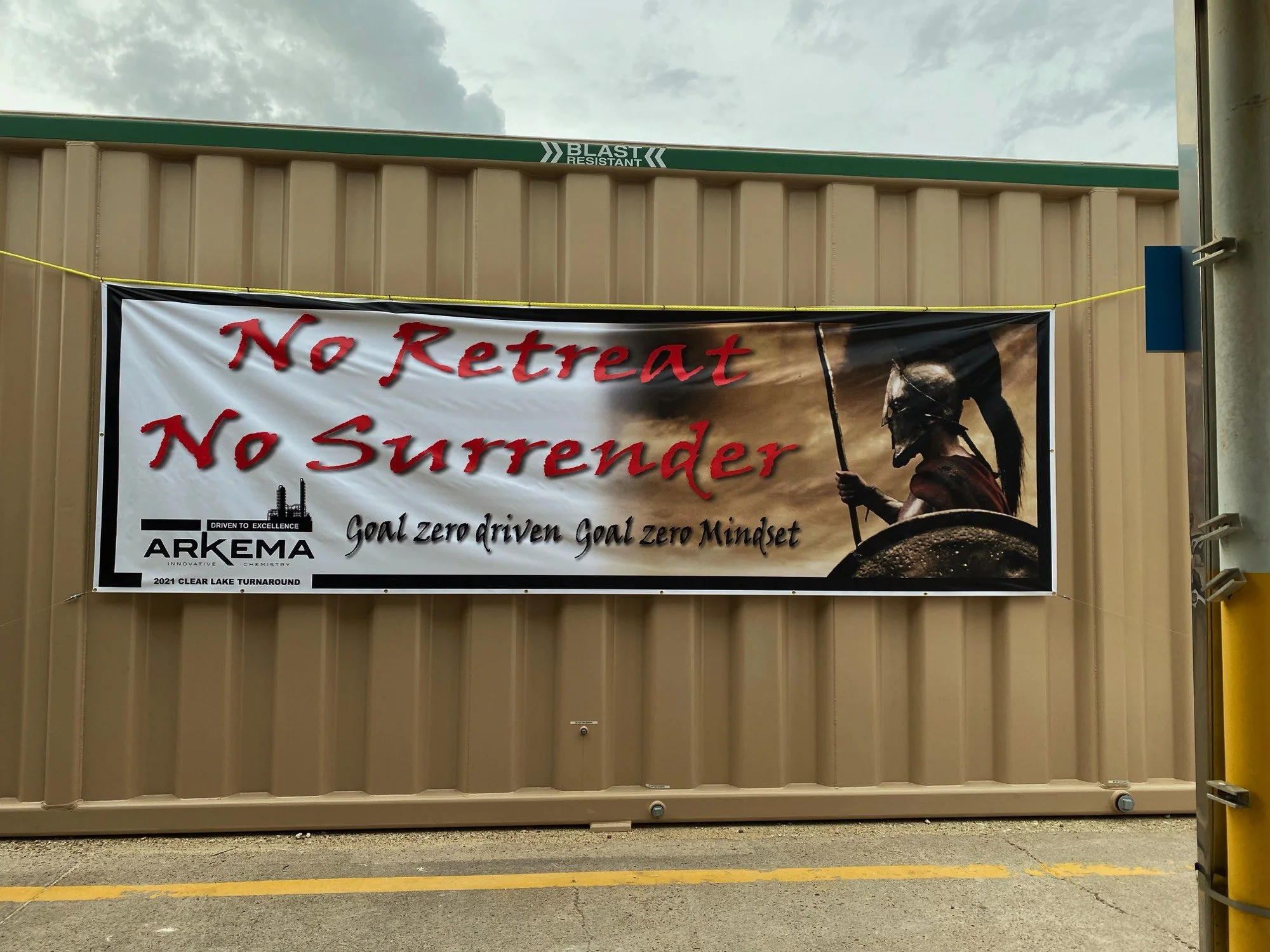A large banner hangs on a beige metal building, displaying the message "No Retreat No Surrender" in red text. The banner includes an image of a warrior wearing a helmet and armor, holding a spear and shield. There is also a logo for Arkema and a slog
