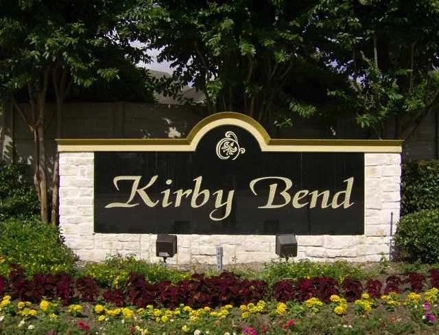 Sign reading 'Kirby Bend' in front of trees and colorful flowers.