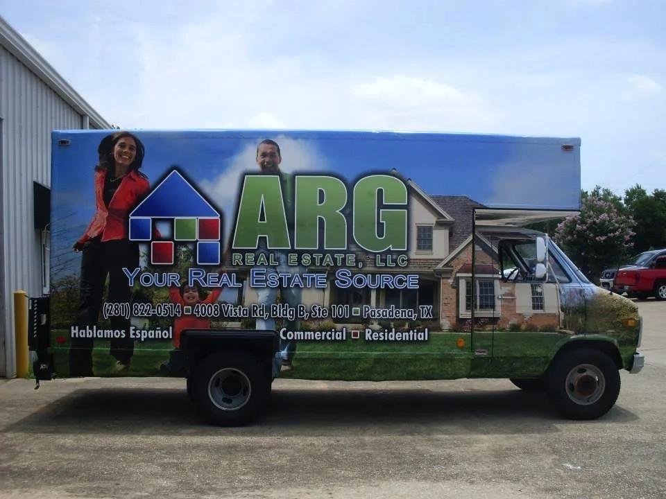 A large truck advertising ARG Real Estate LLC features images of smiling people, a house, and the company's contact information, with text indicating services in both English and Spanish.
