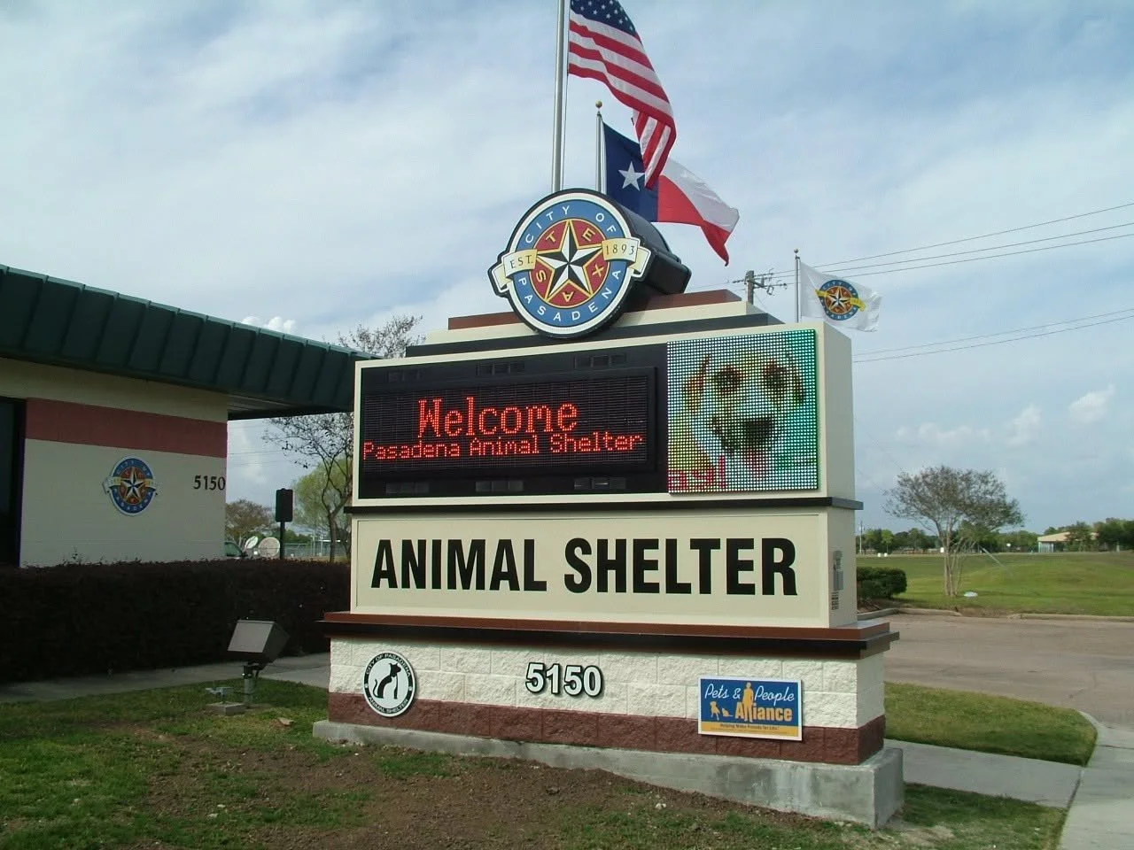 Sign at Pasadena Animal Shelter displaying welcome message with a picture of a dog, flags flying in the background, and the shelter's logo.