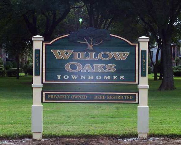 Sign for Willow Oaks Townhomes with a large tree graphic, located in a grassy area with trees in the background.