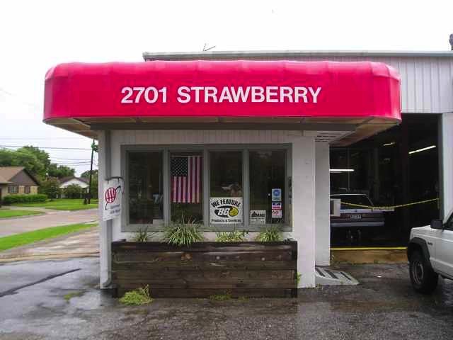 Front of a small storefront with a bright pink awning displaying the address 2701 Strawberry. There are plants in a wooden planter box in front of the window, and an American flag in the window.