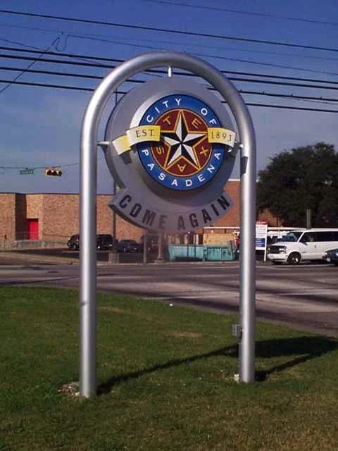 A circular sign on a metal frame with the words "City of Pasadena" and an emblem, with a banner saying "EST 1887", located on a grassy area near a street with cars and buildings in the background.