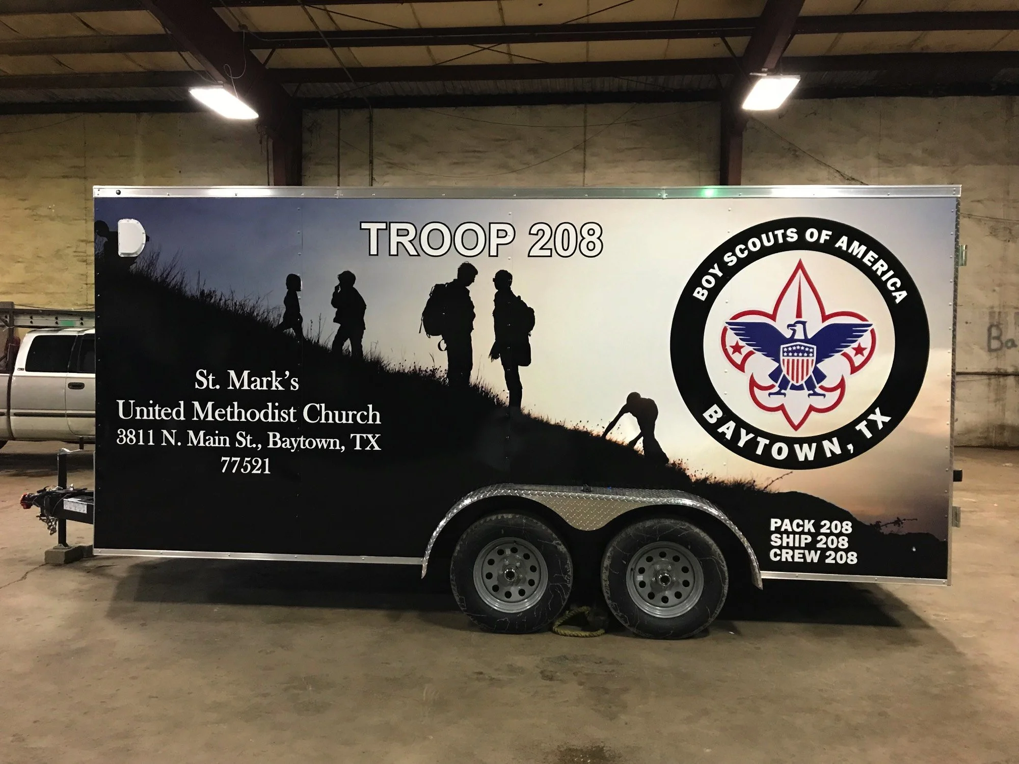 A trailer with Boy Scouts of America insignia and signage for Troop 208, Baytown, Texas. The trailer features silhouettes of scouts hiking and text for the Scout group and their church, located inside a building with an industrial ceiling and fluorescent lights.