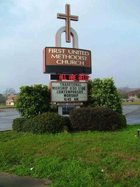 Signboard of First United Methodist Church with a cross on top, displaying service times and a welcome message, surrounded by greenery.