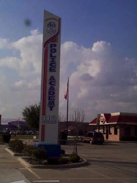 Sign for California Polytechnic State University, featuring the university emblem and name, located outside on a sunny day with a flagpole and parking lot.