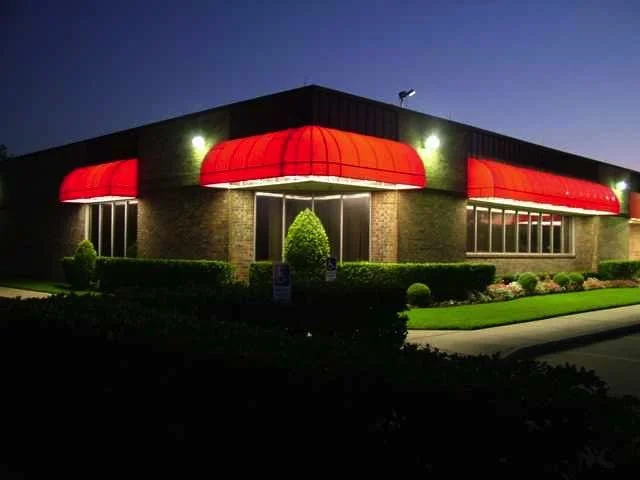 Night-time exterior of a building with red awnings and bright lights, surrounded by manicured bushes and grass.