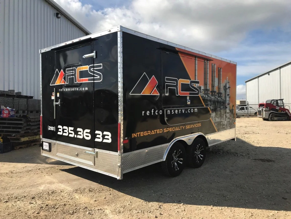Black enclosed trailer with company logo and contact information, parked on gravel ground outside industrial buildings, with some machinery and vehicles in the background.