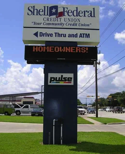 Sign for Shell Federal Credit Union with drive-thru and ATM, digital message displaying 'HOMEOWNERS!', and a parking lot with cars in the background.