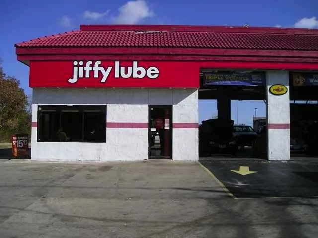 Jiffy Lube automotive service center with a red roof, white walls, and a large sign reading 'jiffy lube' on the front. The entrance is in the center, and there is a driveway leading into the service bay on the right.