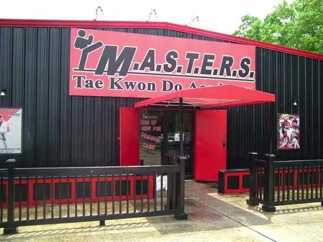 Exterior of a martial arts school called 'YMASTERS Tae Kwon Do Academy' with a red awning over the entrance, black fencing, and posters on the building.
