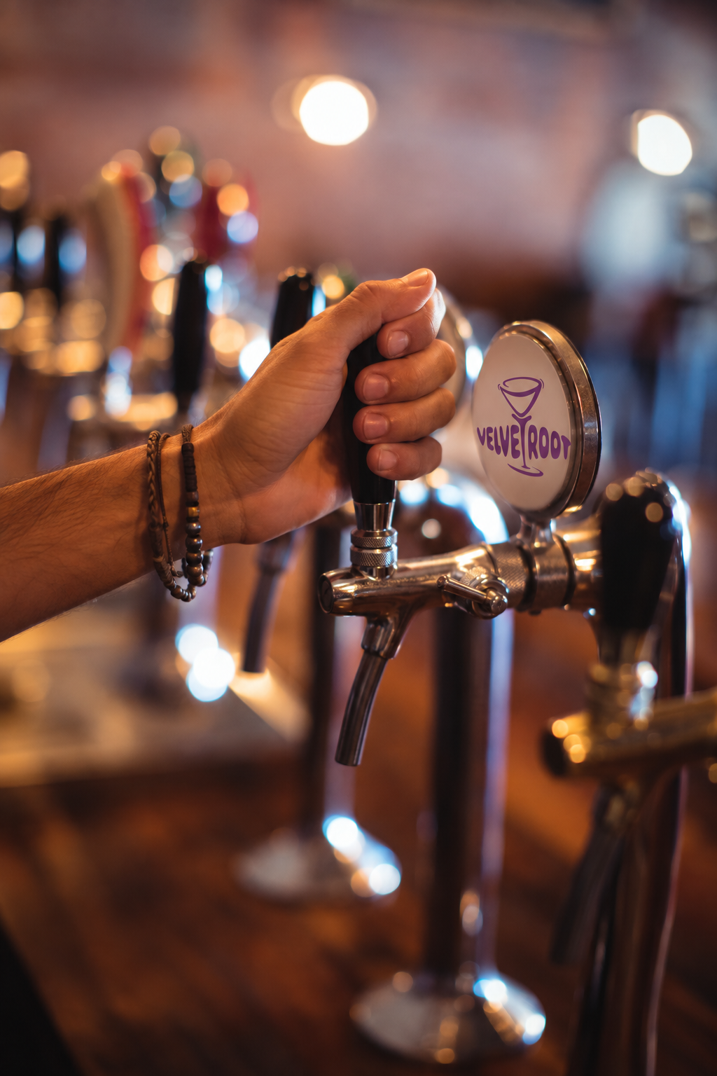 A bartender's hand is pulling a tap handle on a cocktail tap system with a logo labeled 'Velvet Root', with blurred background of cocktail taps and warm lighting in a bar.