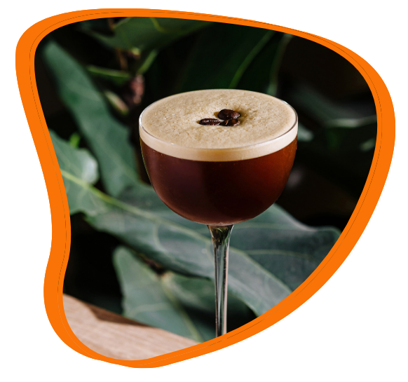 An espresso martini, with a layer of foam and coffee beans on top, set against green leaves.