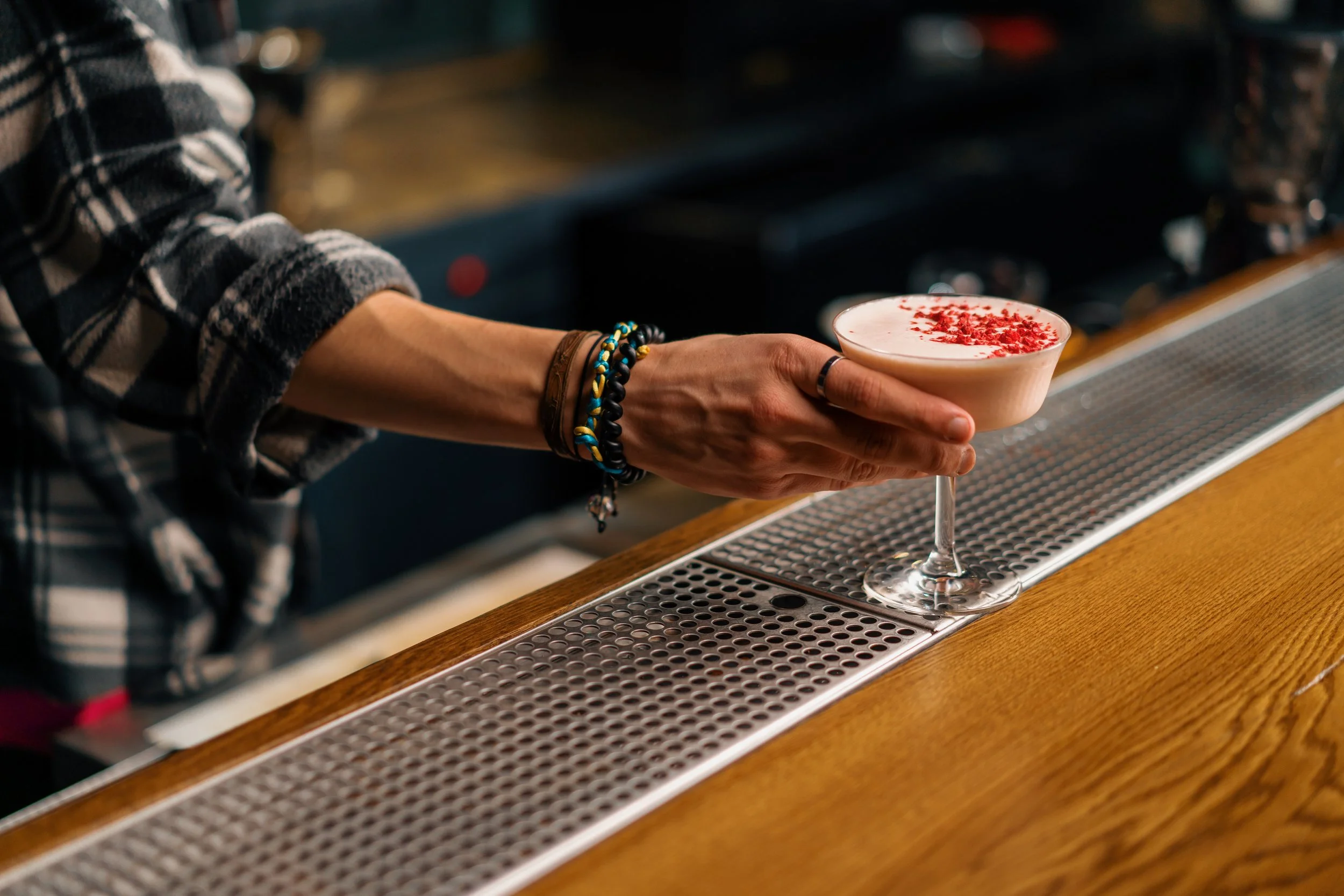 A bartender presenting a stemmed glass filled with a pink cocktail topped with red sprinkles in a bar or coffee shop setting.