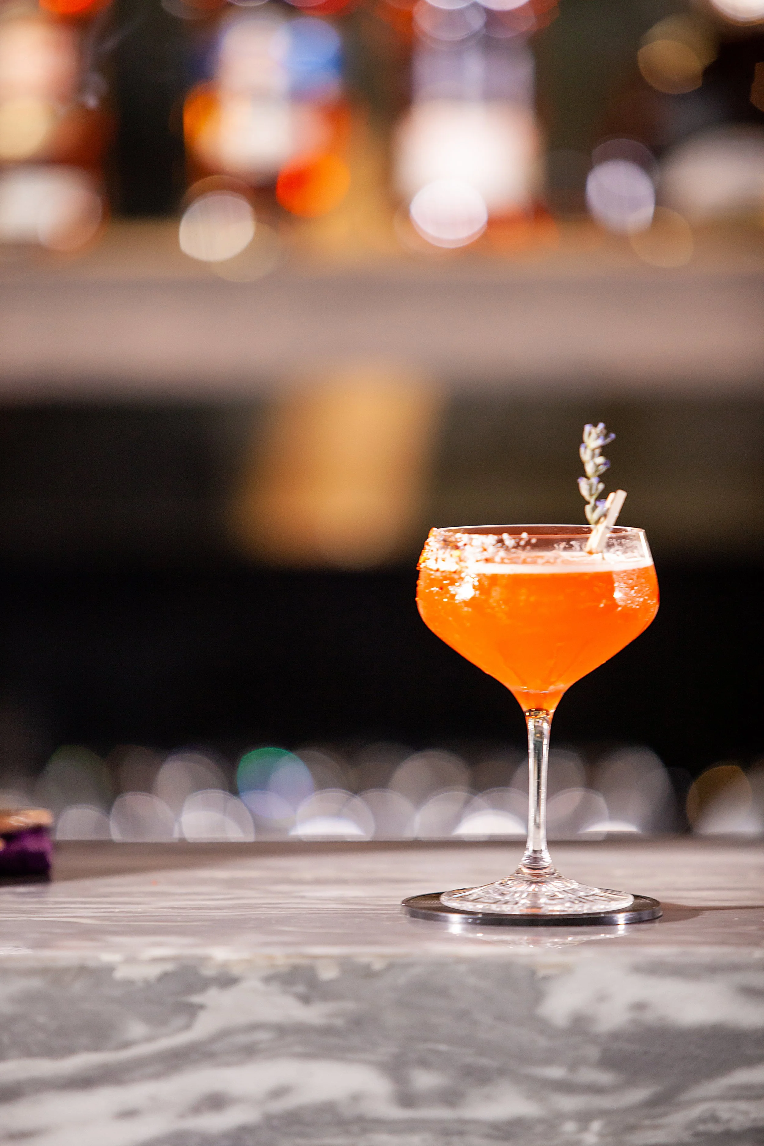 A pink cocktail in a stemmed glass garnished with a sprig of rosemary on a bar counter with blurred city lights in the background.