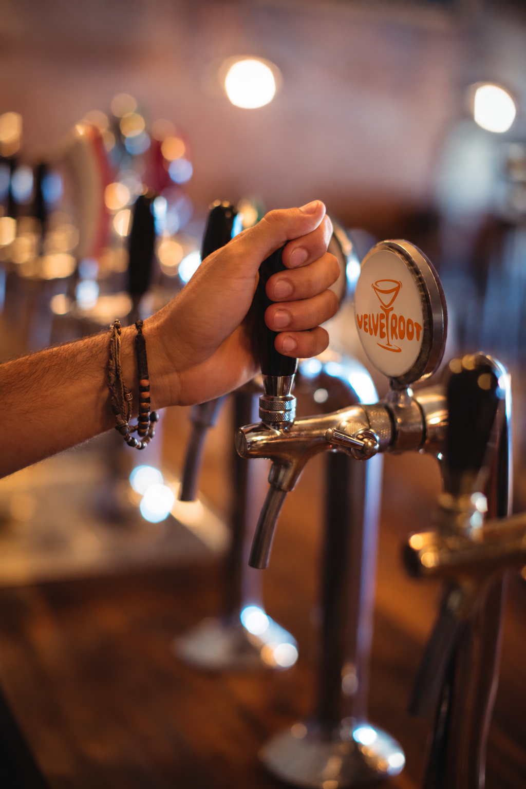Close-up of a person's hand operating a beer tap at a bar. The bar has soft lighting and a blurred background with other beer taps and lights.