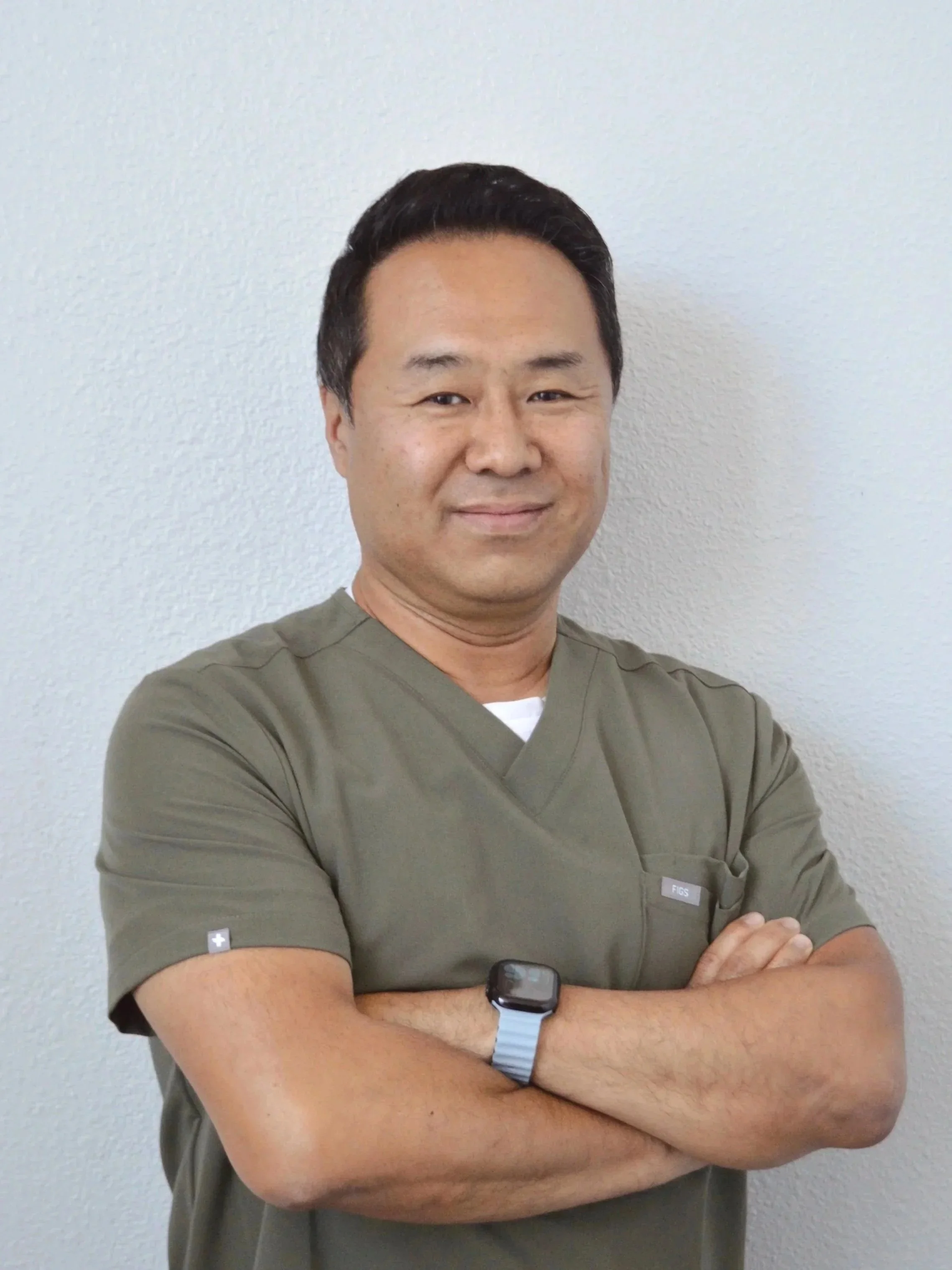 A man in medical scrubs with arms crossed, standing against a plain light-colored wall, wearing a smartwatch.