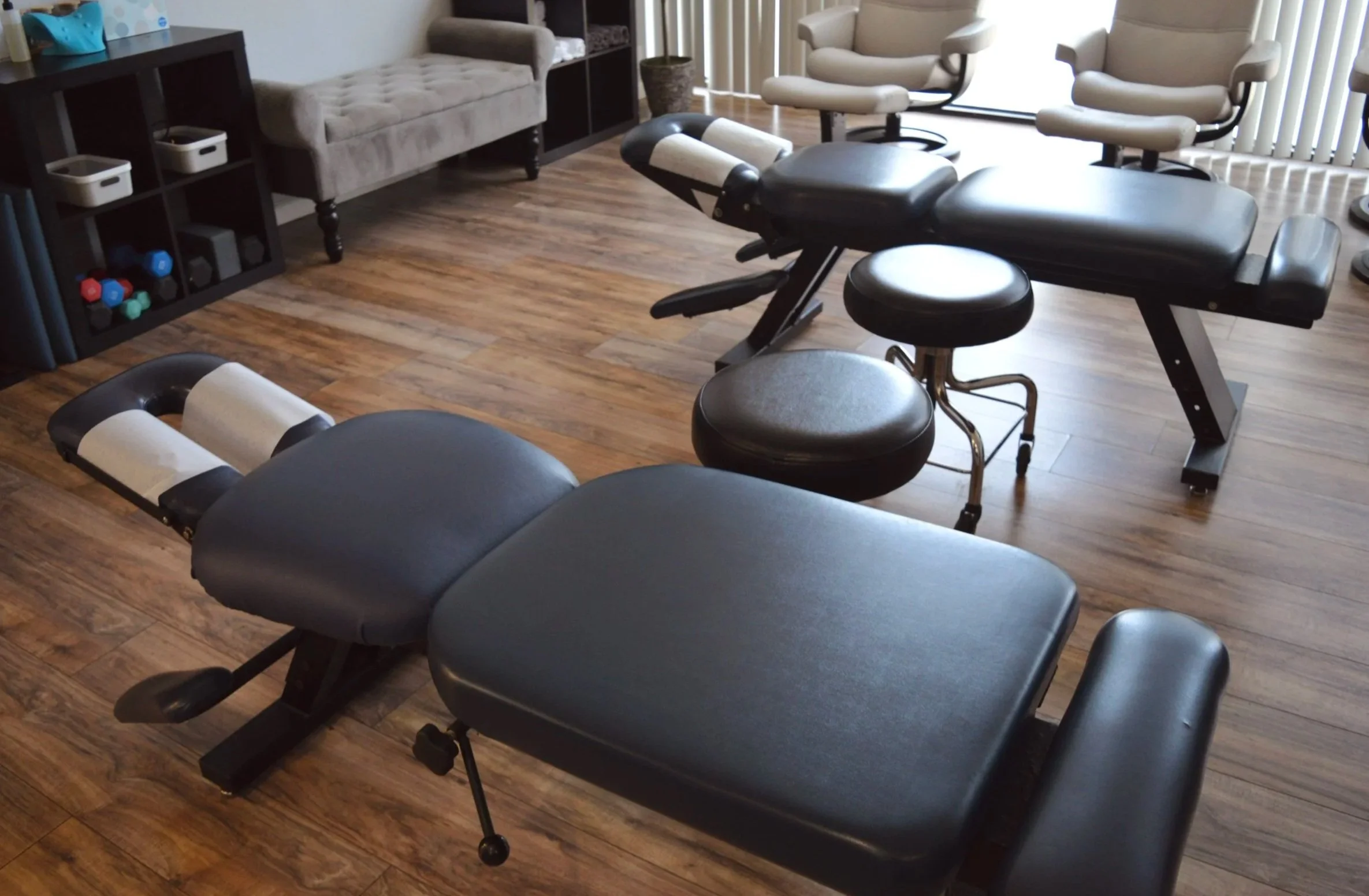 A room with a massage table, two beige lounge chairs, a small stool, a black shelf with gym dumbbells, and a tufted gray sofa, all on a wooden floor with vertical blinds covering windows.