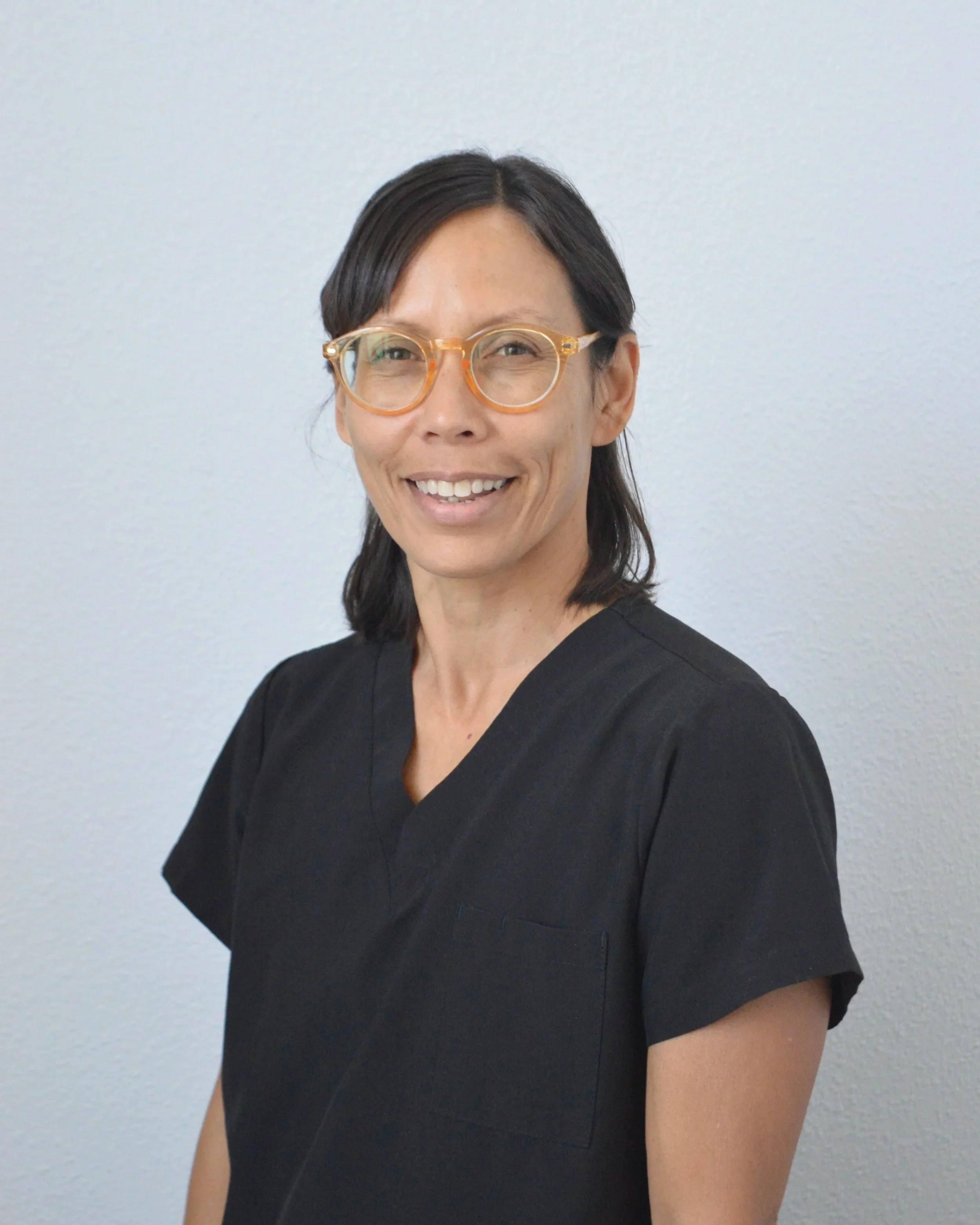 Woman wearing glasses and a black top, smiling, with a plain white wall background.