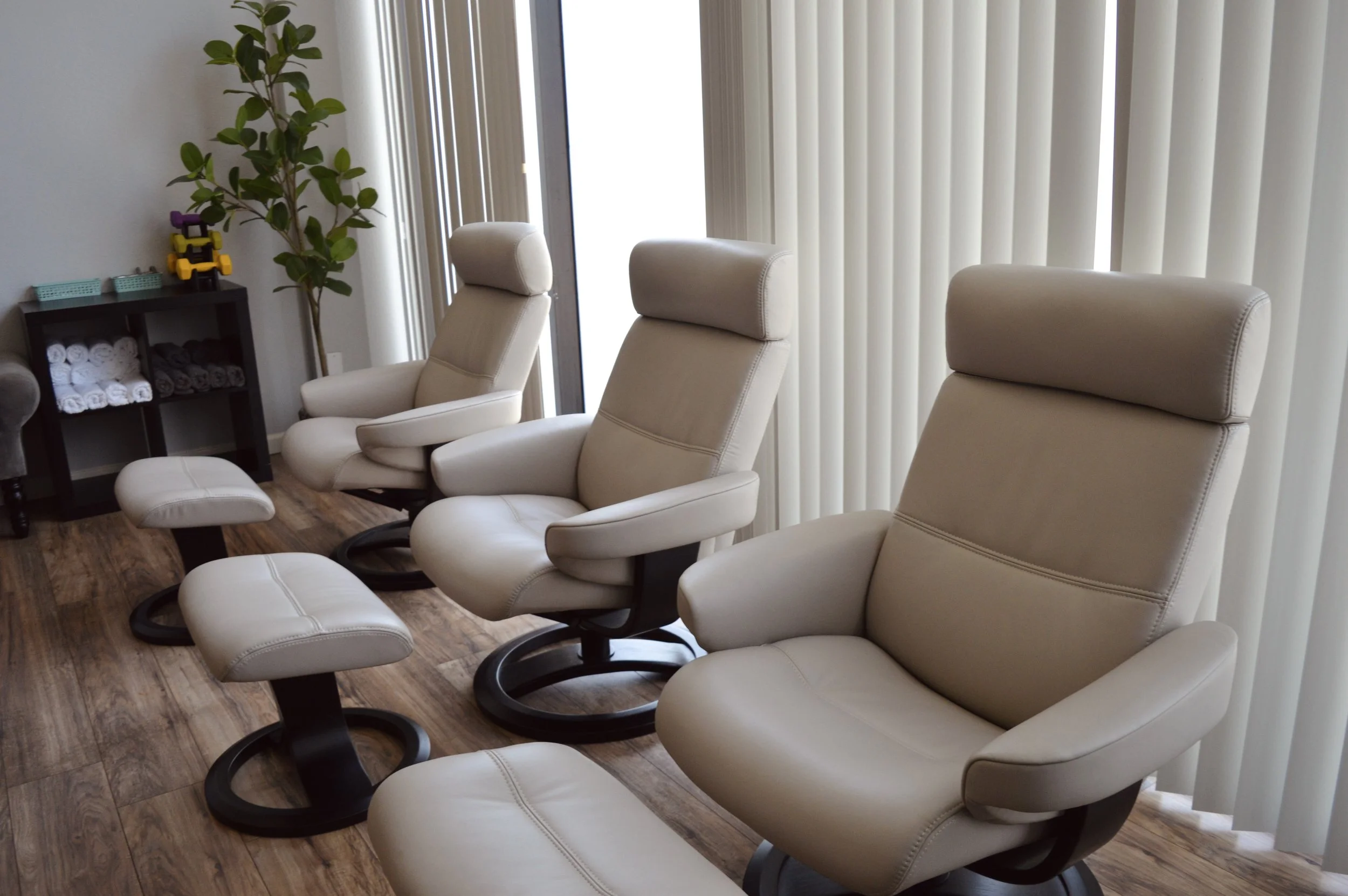 A row of beige massage chairs in a waiting room with wood flooring. Behind the chairs, there is a window with vertical blinds and a small black table holding towels and a decorative sculpture with toy cars.