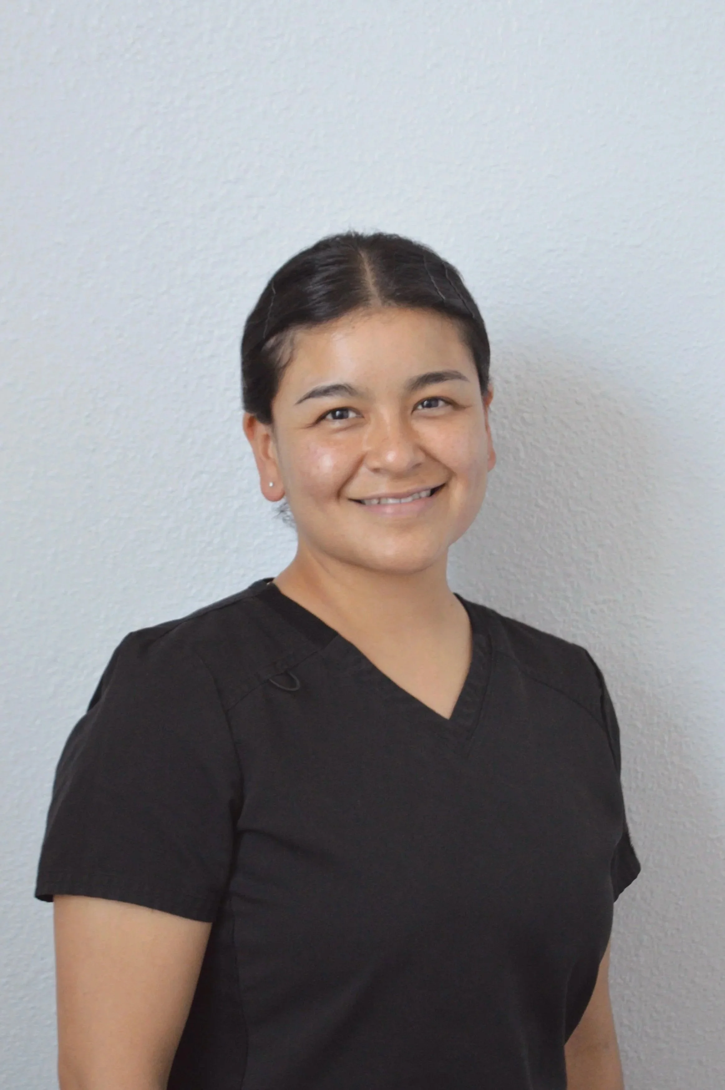 A smiling person with short dark hair wearing a black uniform standing in front of a plain light-colored wall.