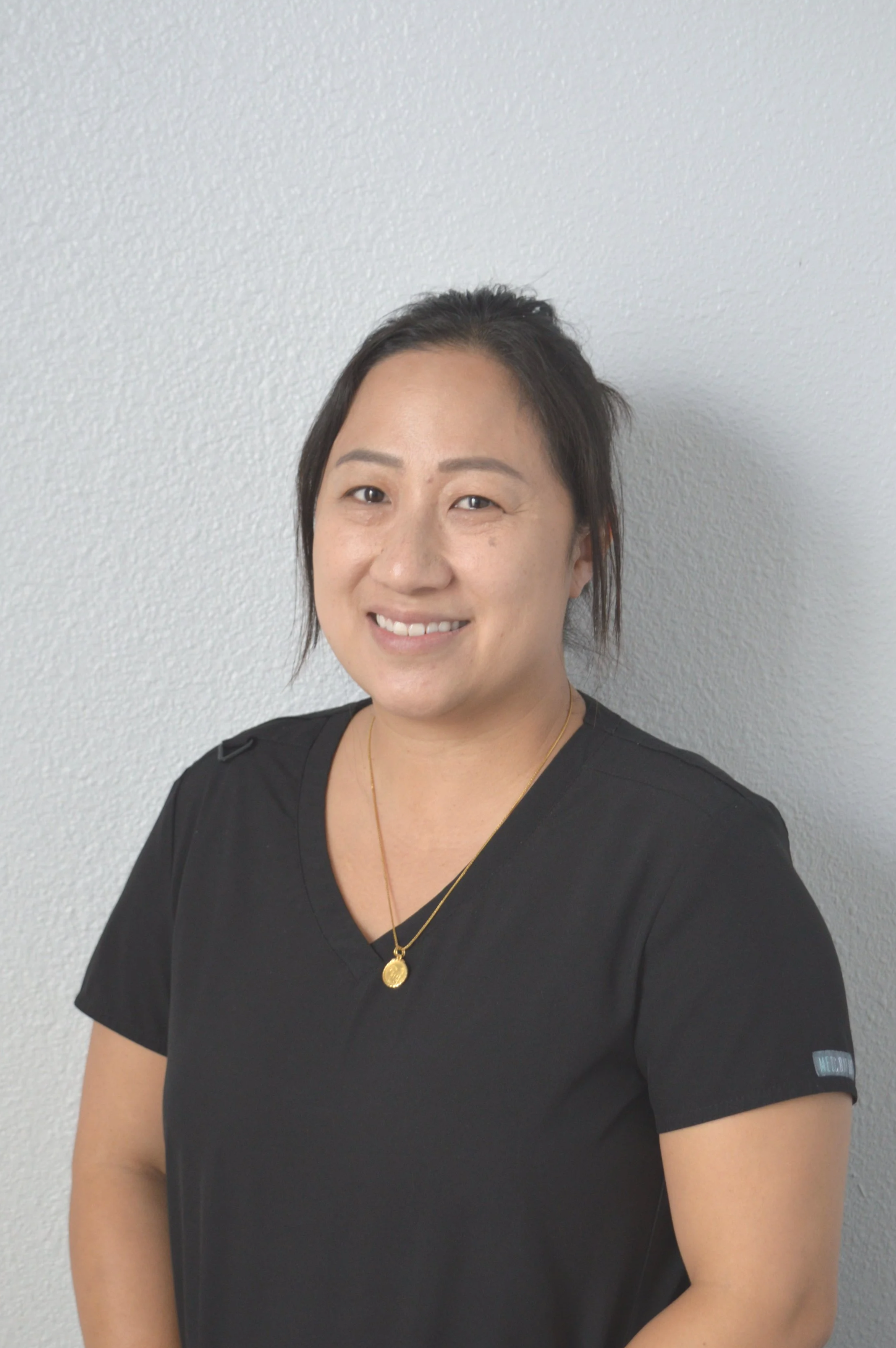 A woman with short dark hair, smiling, wearing a black shirt and gold jewelry, standing against a plain, light-colored textured wall.