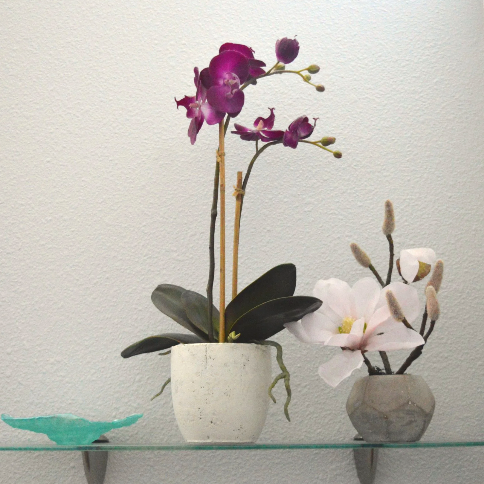 A display of artificial flowers in vases on a glass shelf against a light gray wall. The flowers include purple orchids, white and pink flowers, and green leaves.