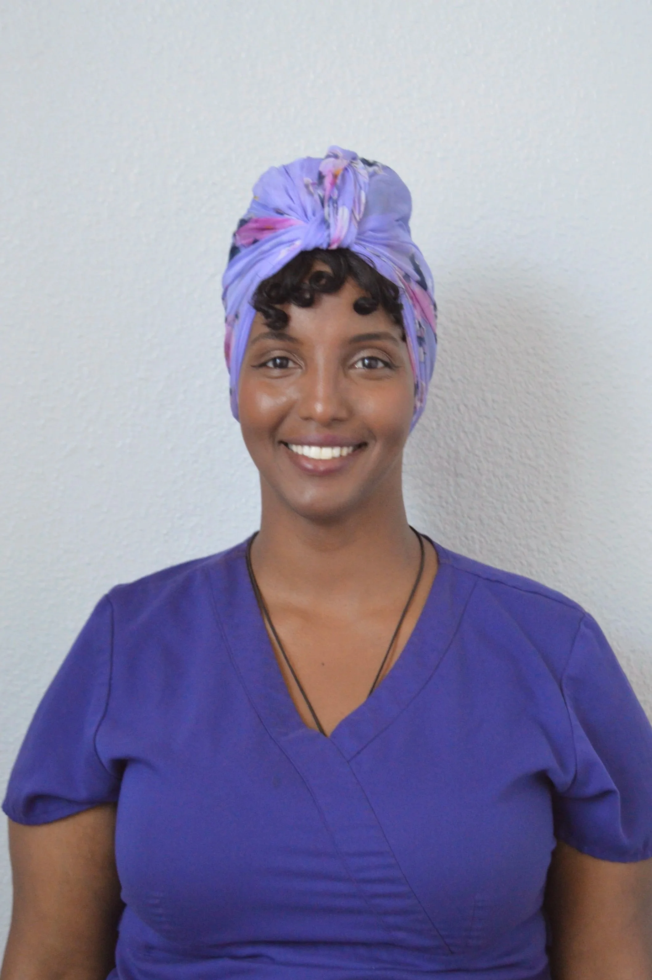 A woman with dark curly hair wearing a blue headscarf and purple scrubs, smiling, against a plain white wall.