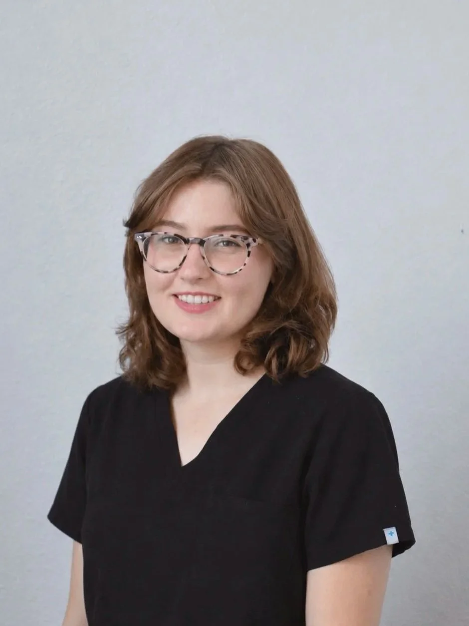 A young woman with shoulder-length wavy brown hair, wearing glasses with a clear and patterned frame, smiling, dressed in a black V-neck shirt, standing against a plain light gray background.