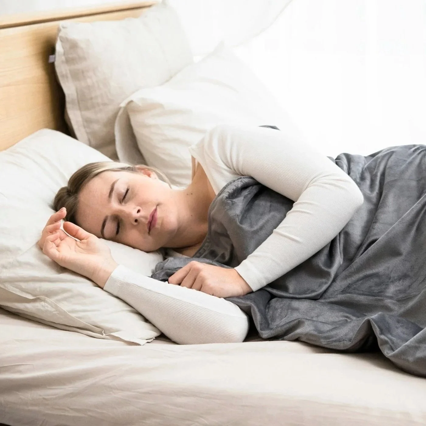 A woman sleeping peacefully in bed with white pillows and sheets, wearing a long-sleeved white shirt and laying on her side.