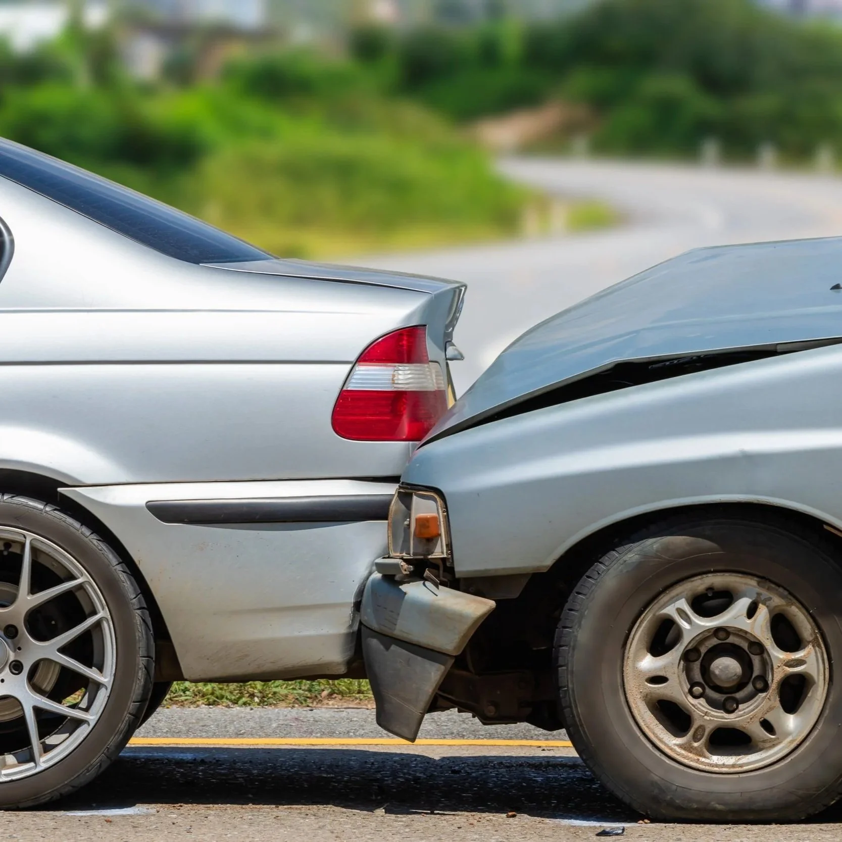 Two cars have been involved in a rear-end collision, with the front of the darker colored car striking the rear bumper of the lighter colored car, causing visible damage.