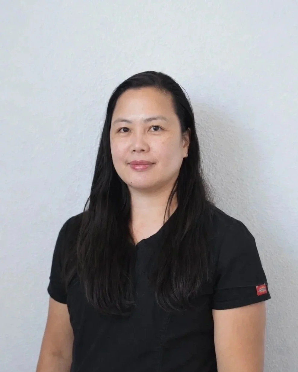 A woman with long dark hair wearing a black top standing against a plain light gray textured wall.