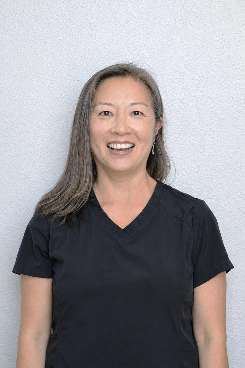 A smiling woman with long gray hair wearing a black medical scrub top standing against a light-colored textured wall.