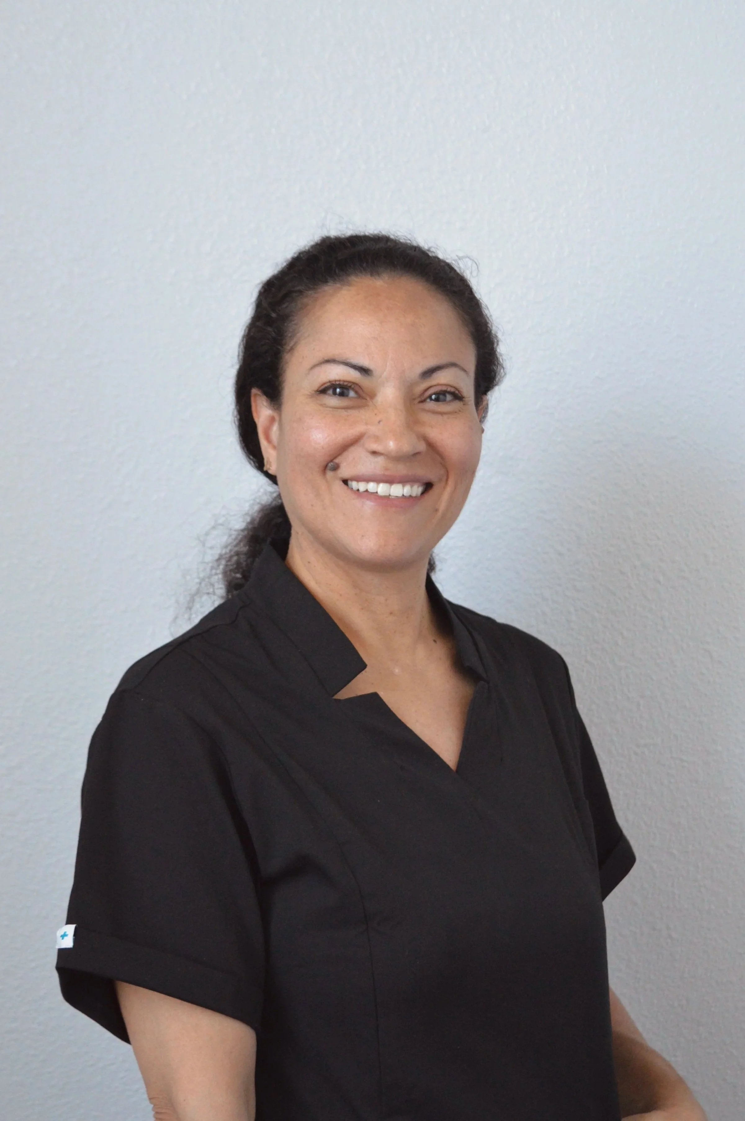 A woman with dark hair tied back in a ponytail, smiling, wearing a black medical scrub top, standing against a plain white wall.