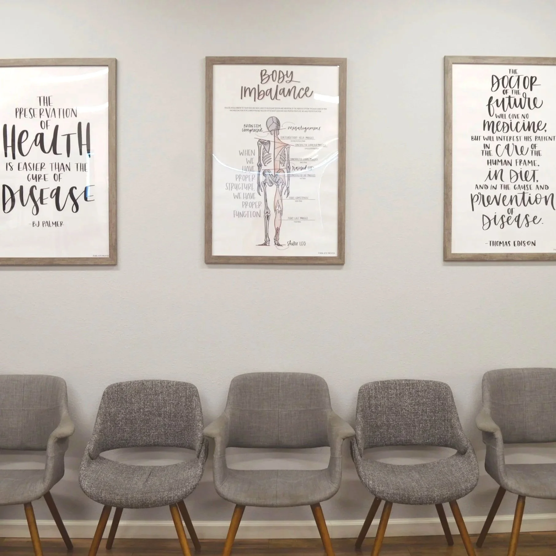 Five gray upholstered chairs with wooden legs arranged in a row against a white wall. Above the chairs are three framed posters with quotes about health, body imbalance, and medicine.