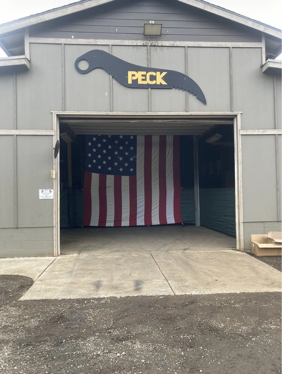 The entrance of Peckweld building with a sign shaped like an excavator thumb that says 'PECK' above the door. Inside, an American flag hangs, filling the opening.