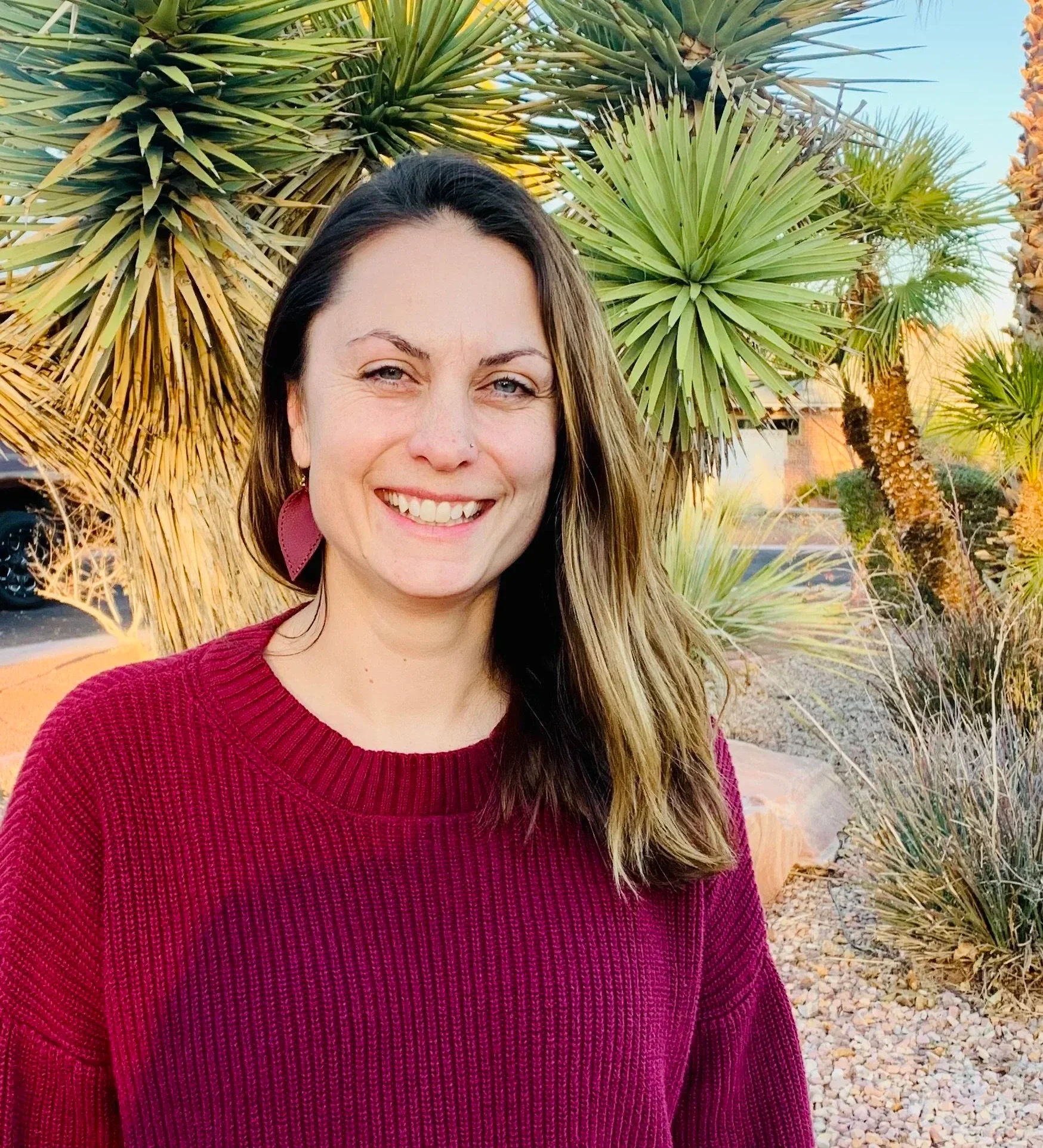 Close-up of a woman with long brown hair, wearing a maroon sweater, smiling outdoors with desert plants in the background.