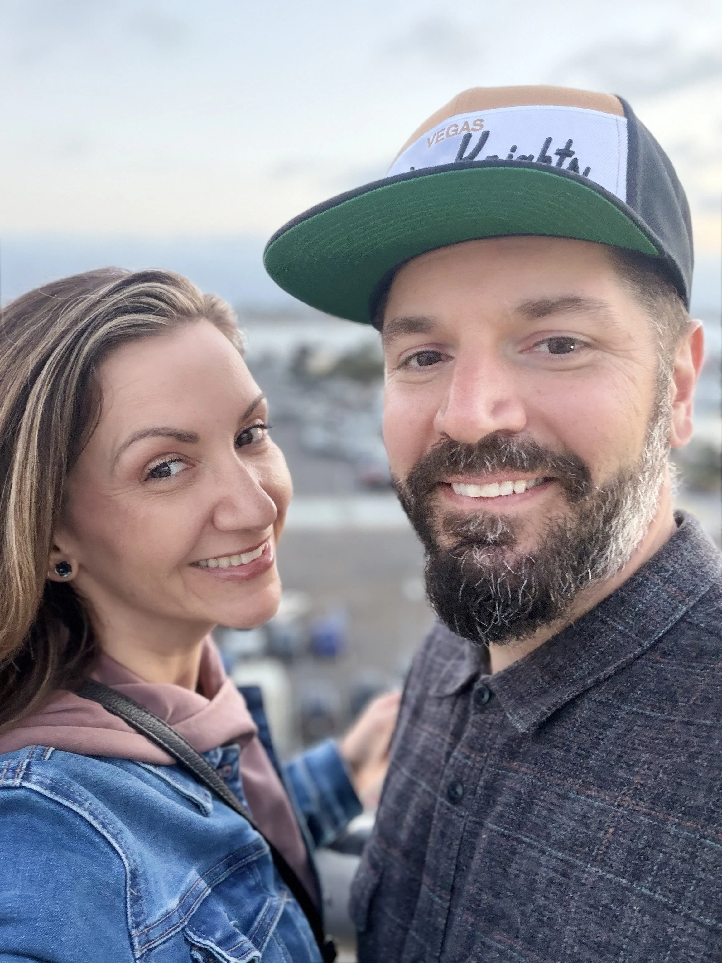 A close-up selfie of a smiling woman and a man. The woman has long, light brown hair and is wearing a denim jacket and pink hoodie. The man has a beard and mustache, wearing a black and tan snapback hat with a patch saying 'Vegas Night' and a dark gray plaid blazer. They are outdoors, with a blurred background of a coastal or ocean scene during sunset or dusk.