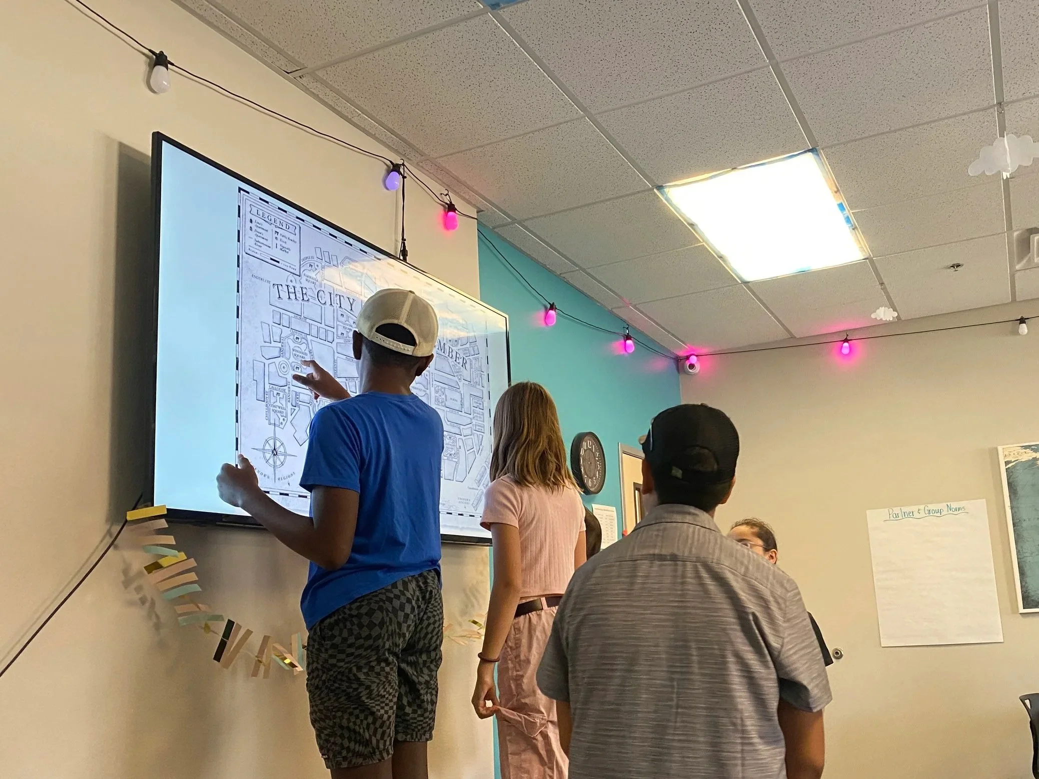 A classroom with students standing in front of a large screen displaying a map. The ceiling has string lights with pink and purple bulbs and cloud-shaped decorations. The students are facing the screen, and some have their backs to the camera.