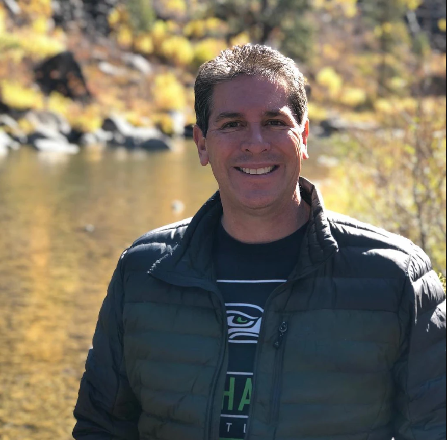 A smiling man outdoors near a river with autumn-colored trees in the background.