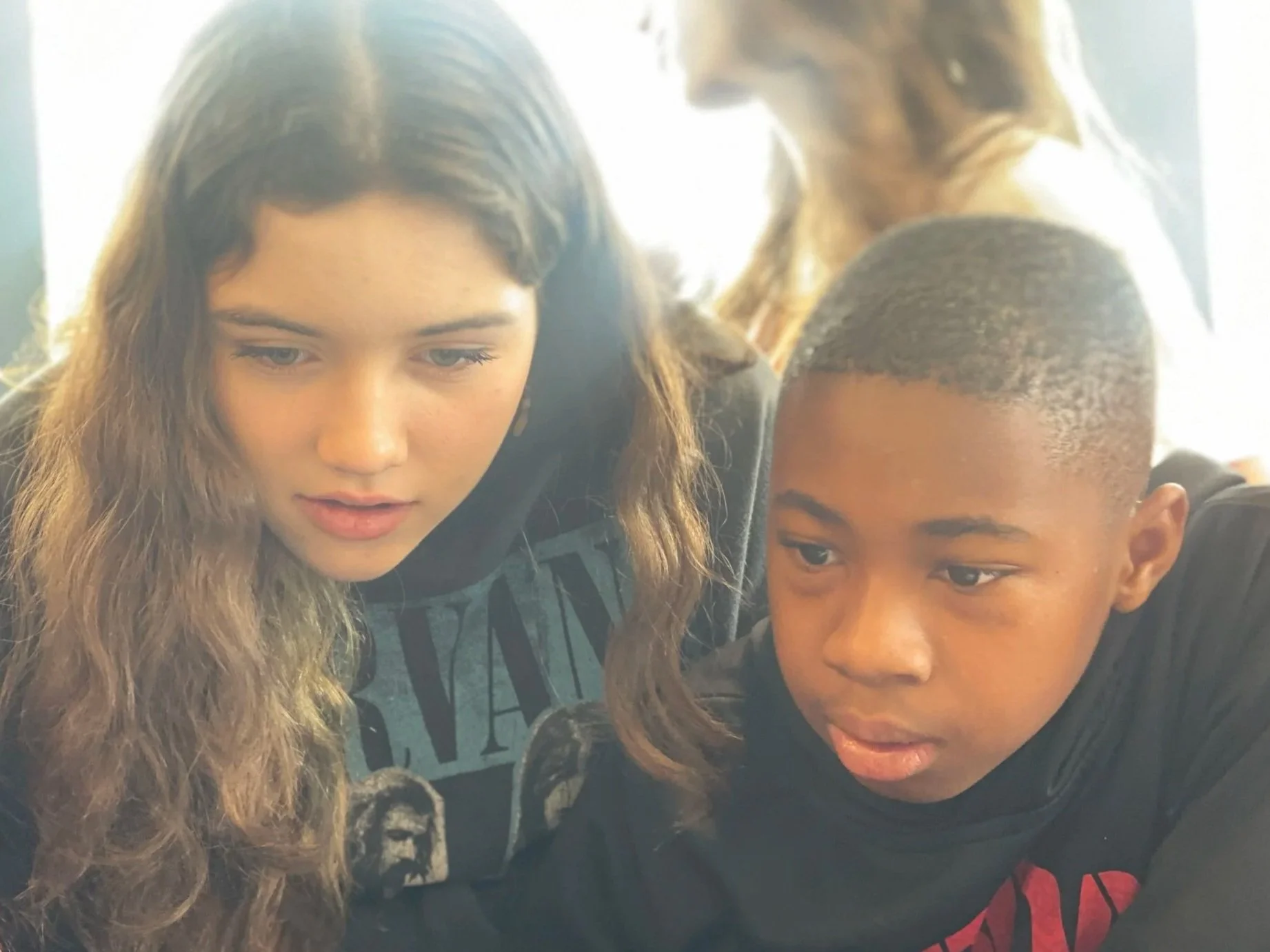 Two young people, a girl with long wavy hair and a boy with short curly hair, are closely engaged with their heads tilted downward, appearing to look at something together.