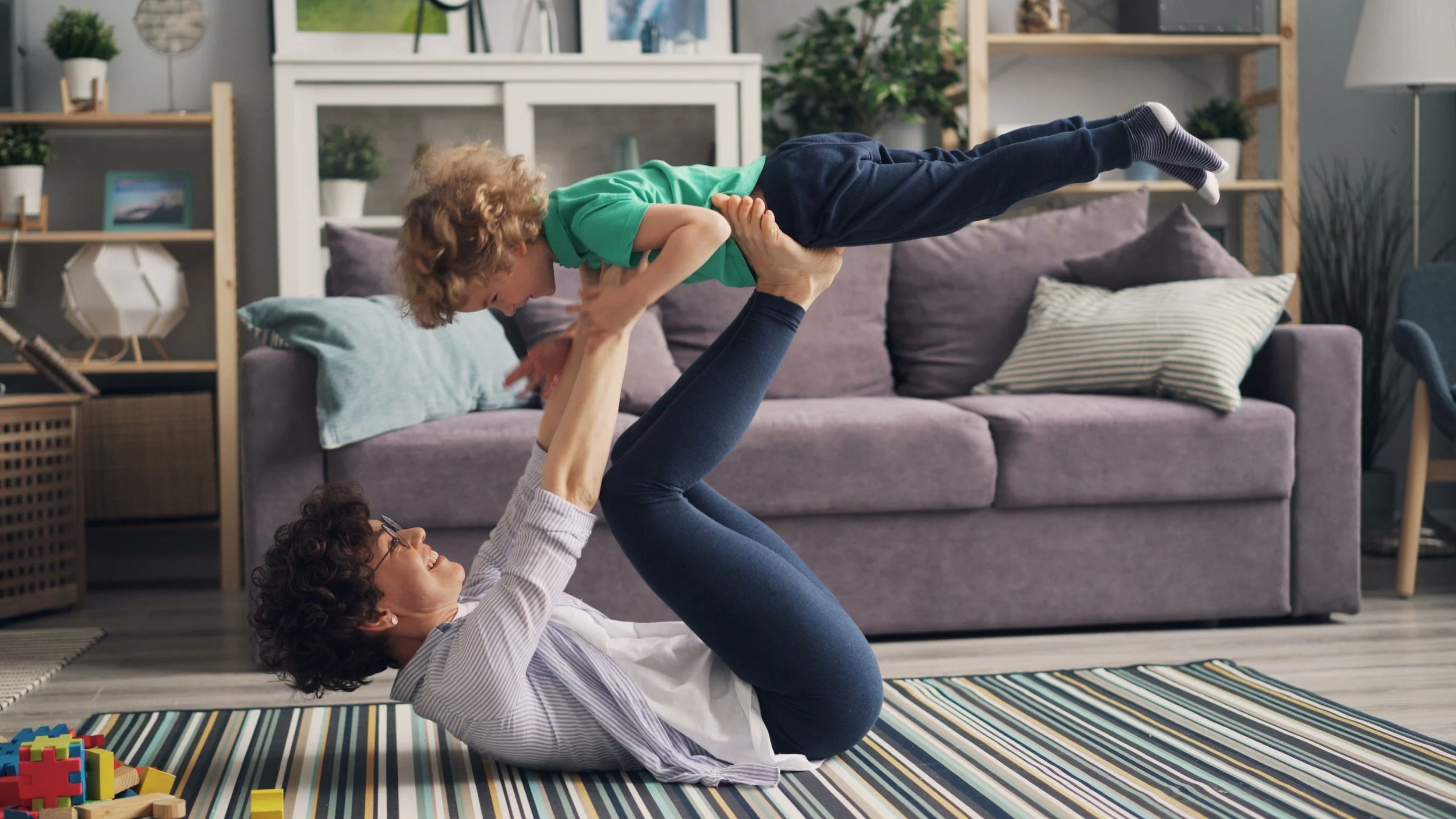A woman lying on the floor with her legs raised and holding a young child in the air above her. The woman is smiling and wearing glasses, while the child has curly hair and is wearing a green shirt. They are playing in a living room with a gray sofa, pillows, shelves, and decorative plants.