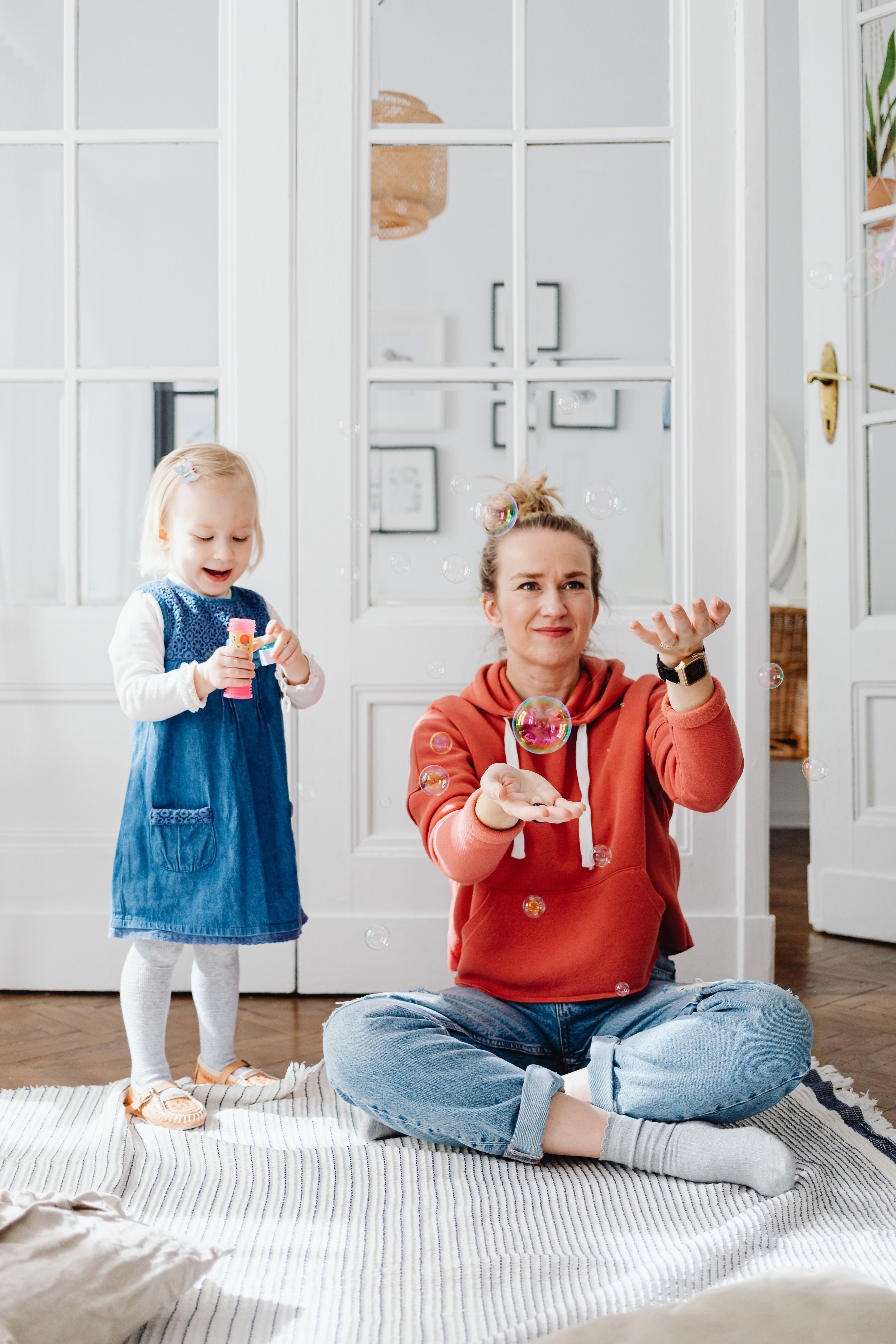 A woman and a young girl playing with bubbles inside a home, with the woman sitting on a striped rug and the girl standing nearby holding a bubble toy.