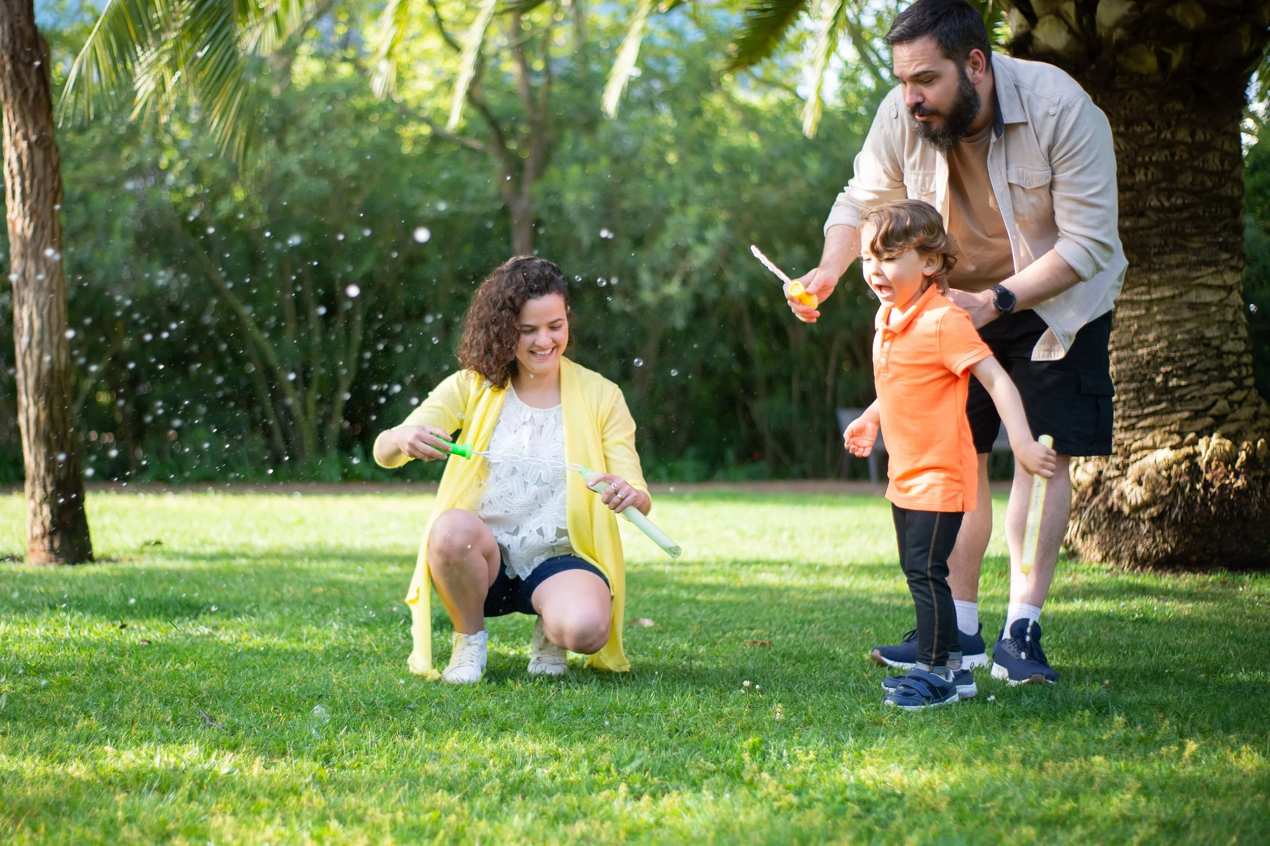 A family of four playing with bubbles outdoors in a park with green trees. The mother is kneeling on the grass, blowing bubbles. The father, standing near a large palm tree, is holding a bubble wand. A young boy in an orange shirt is excitedly watching the bubbles, and a woman in a yellow jacket is smiling while holding a bubble gun.