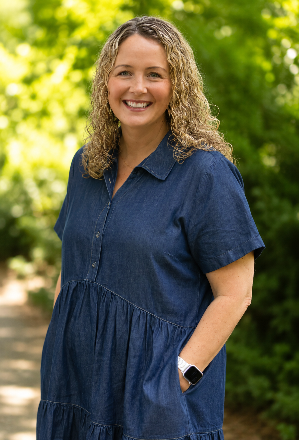 A smiling woman with curly blonde hair wearing a denim dress, standing outdoors with greenery in the background.