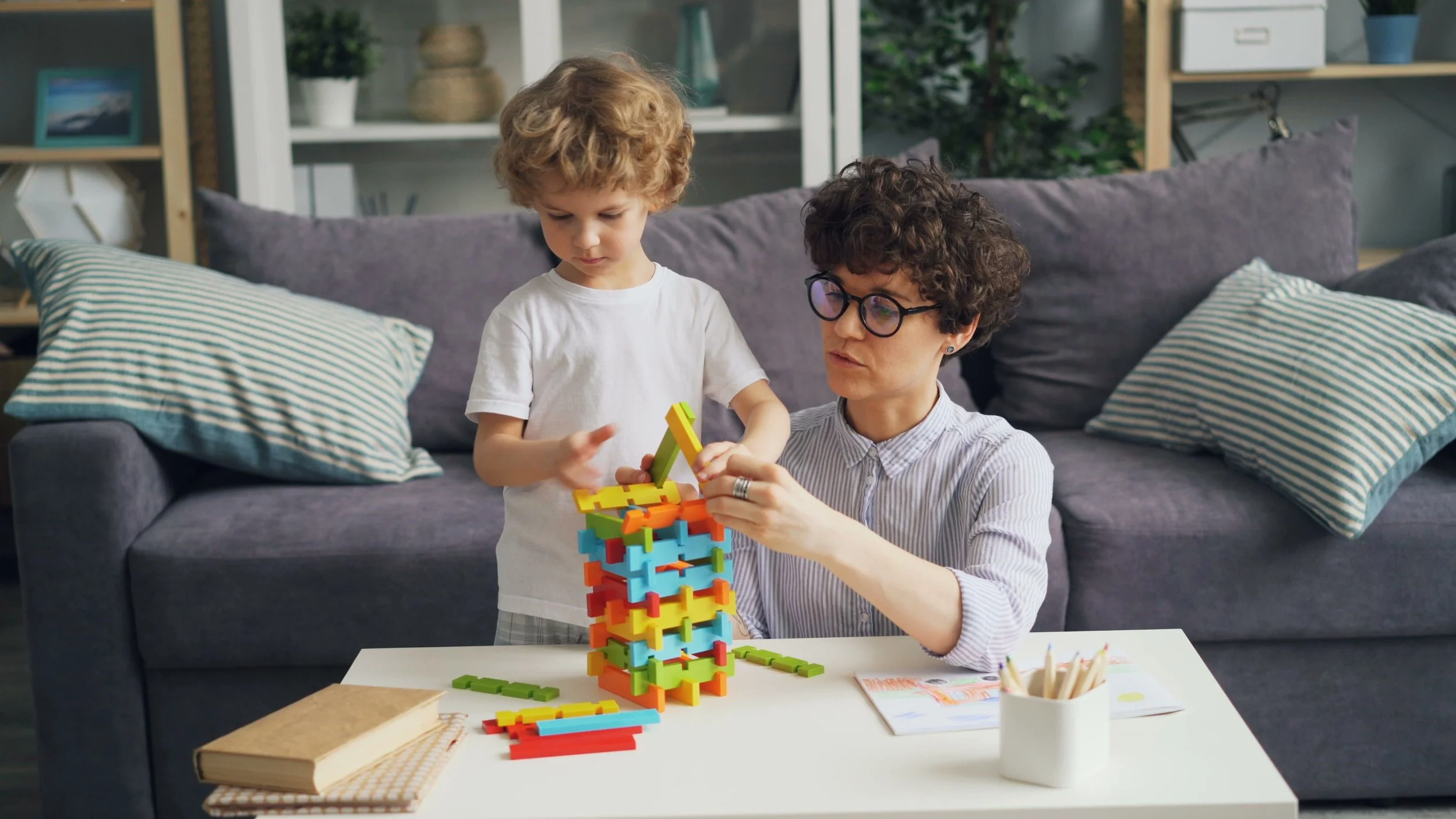 A woman with short curly hair, glasses, and a striped shirt is sitting at a table, playing a colorful tower-building game with a young boy with curly blond hair wearing a white t-shirt. They are indoors with a gray couch and shelves in the background.