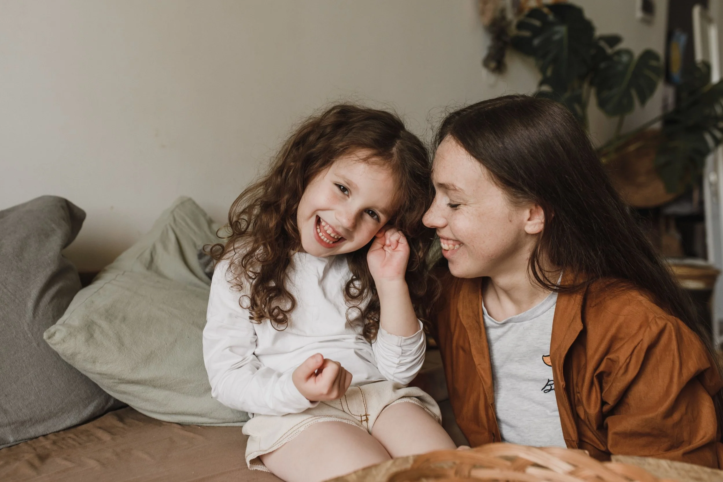 A young girl with curly brown hair smiling and whispering into an adult woman's ear, who is also smiling. They are sitting on a beige couch with pillows and a large plant in the background.