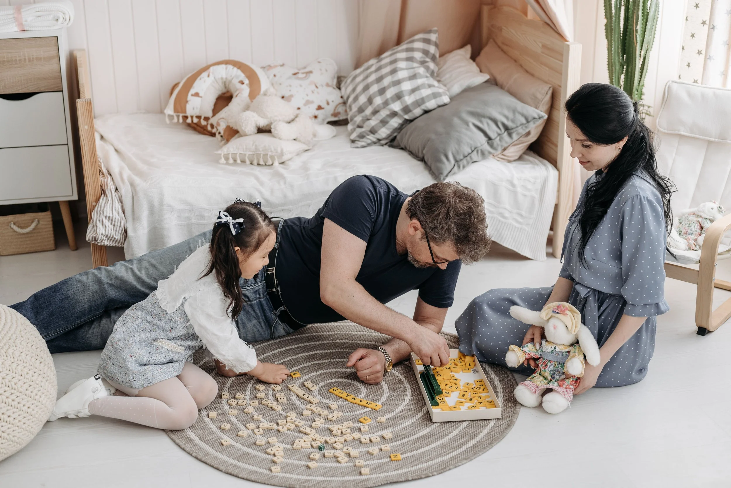 A family is playing Scrabble on a round rug in a bedroom with a bed, pillows, and stuffed animals in the background. The family includes a man, a woman, and a young girl.