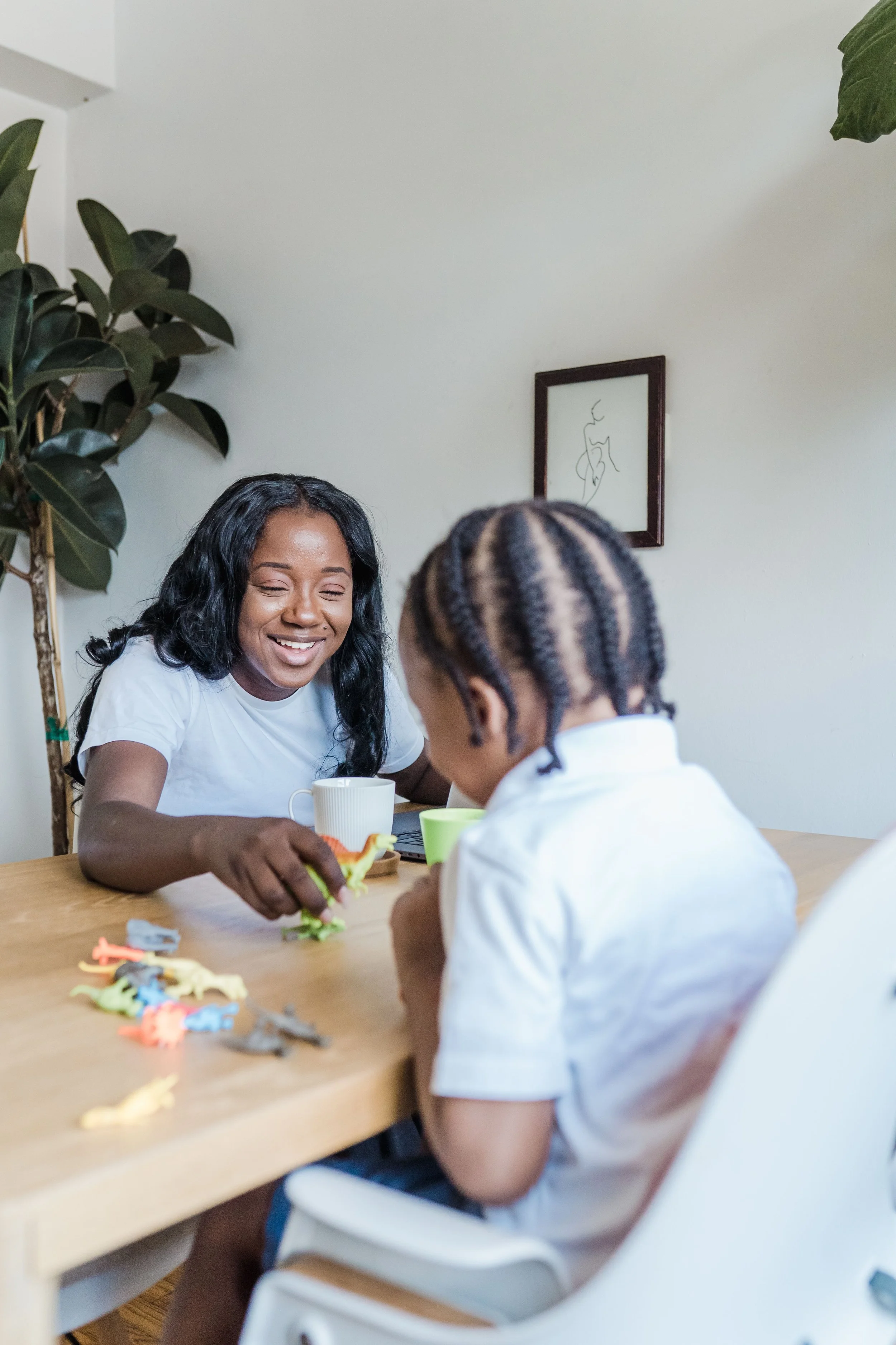 A woman and a child sitting at a wooden table with toys and utensils, smiling and engaging with each other in a bright room.